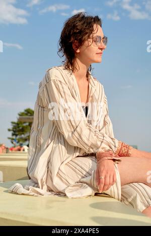 Eine Frau sitzt entspannt im Freien, trägt ein gestreiftes Hemd und eine Sonnenbrille und blickt zur Seite mit einem klaren blauen Himmel im Hintergrund. Stockfoto