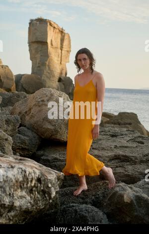 Eine Frau in gelbem Kleid steht auf felsigem Gelände mit dem Meer und einer großen Felsformation im Hintergrund am späten Nachmittag. Stockfoto