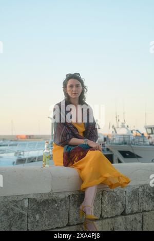 Eine Frau, die in der Abenddämmerung auf einem Steinvorsprung an einem Jachthafen sitzt, ein gelbes Kleid mit einem bunten Schal trägt, während sie einen Drink hält. Stockfoto