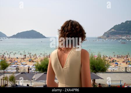 Eine Frau steht auf einem Balkon, mit Blick auf einen überfüllten Strand mit Leuten, die sich im Meer sonnen und schwimmen, mit Hügeln in der Ferne. Stockfoto