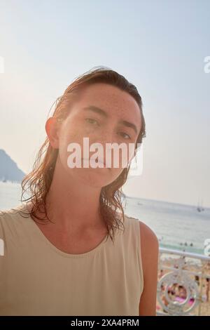 Eine junge Frau mit Sommersprossen steht vor einem sonnigen Strand-Hintergrund, ihre Haare wurden zur Seite gefegt, und das Sonnenlicht strahlt ein sanftes Licht auf ihr Gesicht. Stockfoto