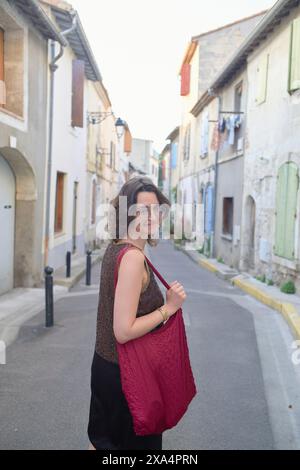 Eine junge Frau mit schulterlangen Haaren lächelt und blickt über die Schulter, während sie eine enge Straße entlang geht, die von traditionellen Häusern im europäischen Stil gesäumt ist. Stockfoto
