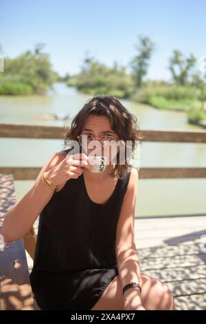 Eine junge Frau sitzt draußen an einem sonnigen Tag, mit einer Sonnenbrille, mit Wasser und Grün im Hintergrund. Stockfoto