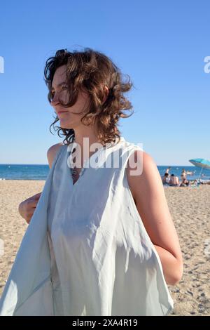 Eine junge Frau steht an einem Sandstrand mit dem Meer im Hintergrund unter einem klaren blauen Himmel, trägt ein helles, fließendes Kleid, und ihre Haare wehen in der Brise. Stockfoto