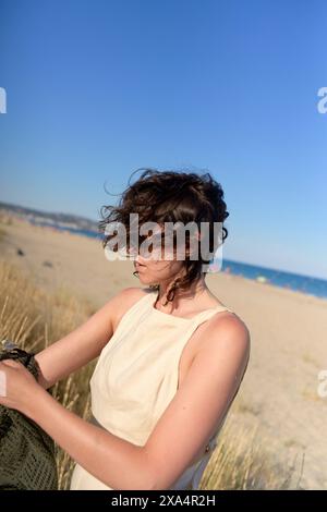 Eine Frau mit lockigen Haaren, die ein ärmelloses Top trägt, ist am Strand, ihr Blick nach unten gerichtet, vielleicht blickt sie in ihre Tasche mit dem Meer und dem blauen Himmel im Hintergrund. Stockfoto