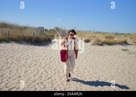 Eine Frau läuft an einem sonnigen Tag auf einem Sandstrand mit Dünen und einem Holzzaun. Stockfoto