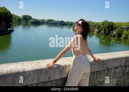 Eine Frau im Sommeroutfit lehnt sich an einem sonnigen Tag auf ein Brückengeländer mit Blick auf einen ruhigen Fluss, mit Bäumen und klarem Himmel im Hintergrund. Stockfoto