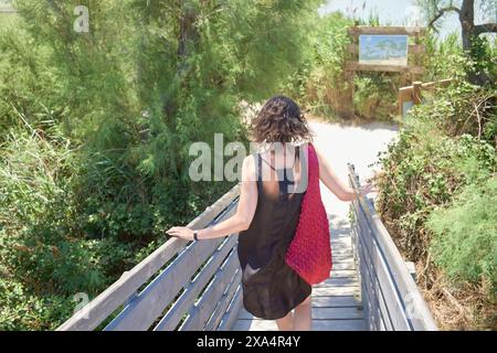 Eine Frau in einem schwarzen Kleid und einer roten Tasche geht einen Holzsteg hinunter zu einem Sandweg, flankiert von grünem Sträucher unter hellem Himmel. Stockfoto