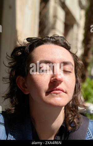 Eine junge Frau mit geschlossenen Augen sonnt sich im Sonnenlicht und erzeugt kontrastreiche Schatten auf ihrem Gesicht. Stockfoto