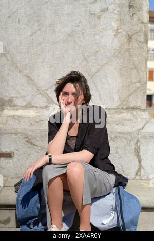 Eine lächelnde Frau mit kurzen dunklen Haaren sitzt lässig draußen auf einer Steinbank und legt ihren Kopf auf ihre Hand. Stockfoto