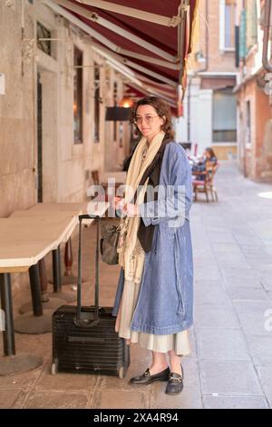 Frau, die in einer malerischen Gasse steht, mit einem Koffer, für Reisen gekleidet. Stockfoto