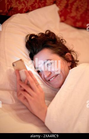 Lächelnde Frau entspannt sich im Bett, während sie ihr Smartphone in einer gemütlichen Umgebung ansieht. Stockfoto