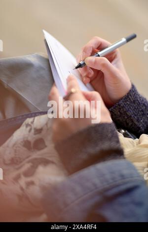 Nahaufnahme der Hände, die mit einem Stift in ein Notizbuch schreiben, auf einem gepolsterten Schoß liegen. Stockfoto