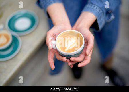 Hände, die eine Keramiktasse mit frisch gebrühtem Kaffee und herzförmiger Latte Art mit mehr Tassen und Untertassen auf einem Tisch im unscharfen Hintergrund wiegen. Stockfoto