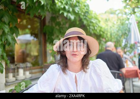 Frau mit Strohhut und Brille, die in einem Café im Freien sitzt, umgeben von Grün. Stockfoto
