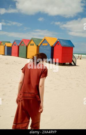 Eine Frau in rotem Outfit steht an einem Sandstrand mit einer Reihe von bunten Strandhütten unter einem bewölkten Himmel im Hintergrund. Stockfoto