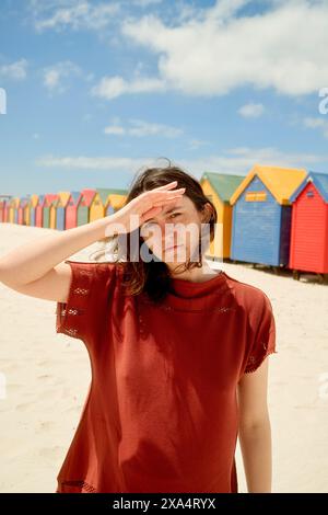 Frau schirmt die Augen vor der Sonne auf einem Sandstrand mit bunten Strandhütten im Hintergrund. Stockfoto