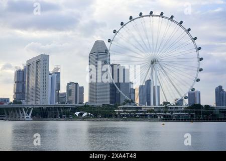 Moderne Skyline der Stadt mit hoch aufragenden Wolkenkratzern und einem großen Aussichtsrad am Ufer unter einem bewölkten Himmel. Stockfoto