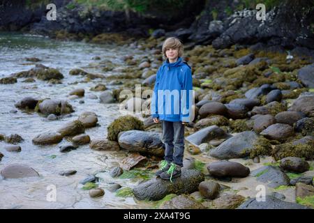 Junge, der an einer felsigen Küste steht, mit Algen und moosbedeckten Felsen um ihn herum. Stockfoto