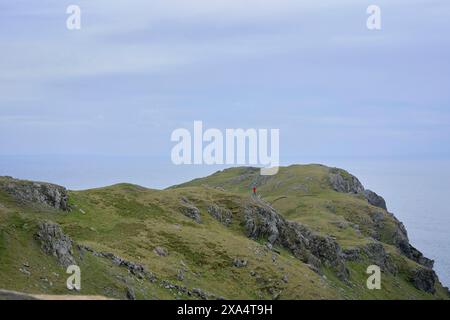 Ein einsamer Wanderer in roter Jacke steht auf einem zerklüfteten Bergweg mit Blick auf das Meer. Stockfoto