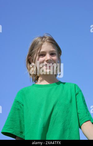Lächelnder Junge in grünem Hemd, der vor einem klaren blauen Himmel steht. Stockfoto