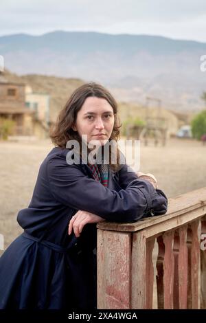 Eine Frau lehnt sich auf einen verwitterten Holzzaun mit einem nachdenklichen Ausdruck vor einem verschwommenen Hintergrund einer ländlichen Landschaft. Stockfoto