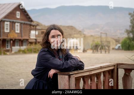 Frau lehnt sich auf einen Holzzaun mit einem rustikalen Haus und Bergen im Hintergrund. Stockfoto