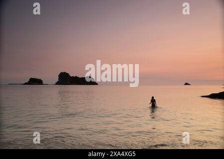 Silhouette einer Person, die bei Sonnenuntergang im Meer steht, mit Felsformationen im Hintergrund. Stockfoto