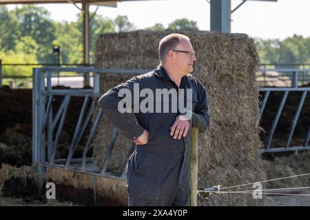 Ein Bauer in einem marineblauen Overall steht selbstbewusst mit den Händen auf der Hüfte, neben Heuballen in einem Bauernhof und blickt in die Ferne. Stockfoto