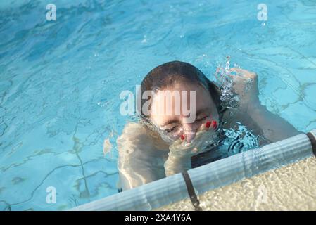 Eine Person taucht aus klarem blauem Poolwasser auf und ergreift mit einer Hand den Rand mit rotem nagellack. Stockfoto