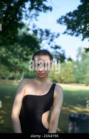 Frau in einem schwarzen ärmellosen Kleid, die an einem sonnigen Tag in einem Park mit Bäumen im Hintergrund steht. Stockfoto