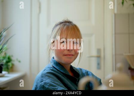 Close-up-Porträt einer lächelnden jungen Frau mit blonden Haaren, die ein Jeanshemd in einer häuslichen Umgebung trägt. Stockfoto