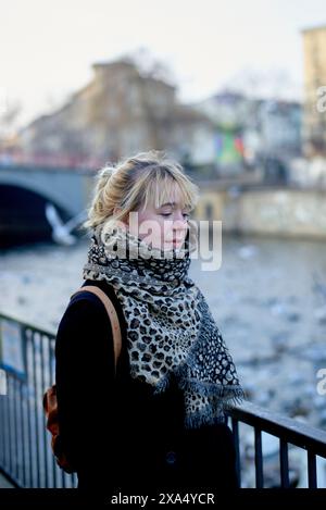 Eine junge Frau mit einem Schal mit Leopardenmuster und einem Rucksack, der an einem Fluss mit Gebäuden und einer Brücke im verschwommenen Hintergrund steht. Stockfoto