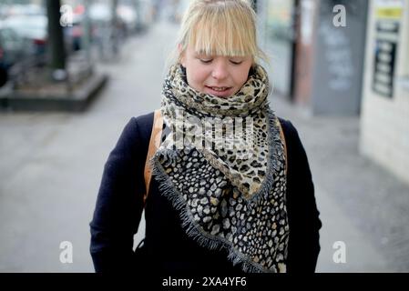 Lächelnde junge Frau mit blonden Pony, die einen Schal mit Leopardenmuster und einen marineblauen Mantel auf einer Stadtstraße trägt. Stockfoto