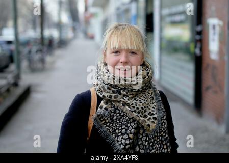 Lächelnde junge Frau mit blonden Haaren und einem gemusterten Schal auf einem Bürgersteig der Stadt. Stockfoto
