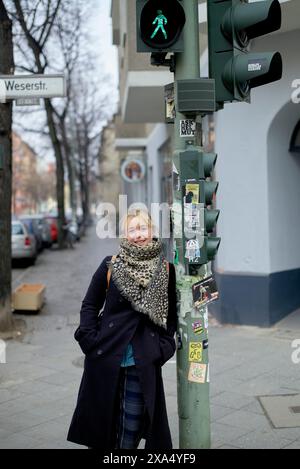 Lächelnde Frau in Winterkleidung, die an einer Ampel mit grünem Fußgängersignal in einer städtischen Umgebung steht. Stockfoto