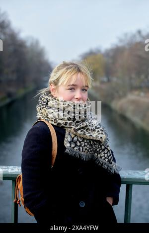 Frau in einem Leopardenschal, lehnt sich an einem Brücken-Geländer mit einem Fluss und Bäumen im Hintergrund an einem nebeligen Tag. Stockfoto