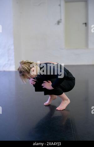 Eine Tänzerin in schwarzer Kleidung steht in einer Hocke auf einem dunklen Tanzstudio-Boden auf ihren Fußballen, mit gebeugtem Kopf und verdecktem Gesicht. Stockfoto