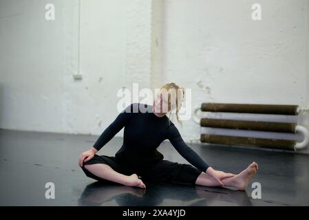Eine Tänzerin in schwarzer Kleidung sitzt auf dem Boden eines Tanzstudios und ruht sich zwischen den Proben aus. Stockfoto