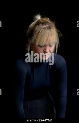 Frau mit blonden Haaren und Pony, die einen schwarzen Rollkragen trägt und vor einem dunklen Hintergrund nach unten blickt. Stockfoto