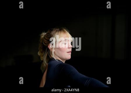 Eine junge Frau mit den Haaren in einem lässigen Brötchen, geschlossenen Augen und hinter dem Rücken gestreckten Armen in einer friedlichen Haltung vor dunklem Hintergrund. Stockfoto