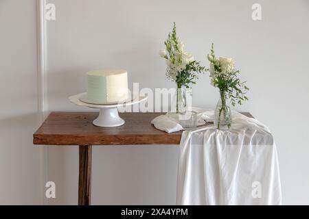 Ein wunderschön dekorierter Kuchen auf einem weißen Ständer sitzt auf einem rustikalen Holztisch, der mit eleganten Vasen aus weißen Blumen und fließenden Stoffen geschmückt ist Stockfoto