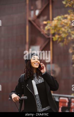 Junge Frau, die mit ihrem Smartphone spricht, während sie an einem Herbsttag in einer Stadtstraße mit einem elektrischen Roller steht. Stockfoto