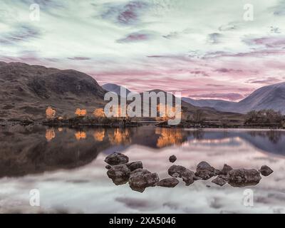 Ruhiger Sonnenuntergang über einem spiegelähnlichen See mit Felsen im Vordergrund und Bergen im Hintergrund unter einem bunten Himmel. Stockfoto