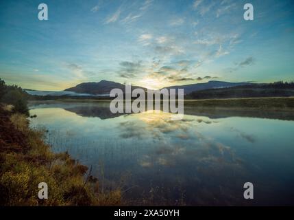 Sonnenaufgang über einem ruhigen See mit Reflexen von Wolken und Hügeln auf dem Wasser, umgeben von bewaldeter Landschaft. Stockfoto