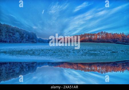 Dämmerungsfarben decken eine ruhige Landschaft mit einem glasklaren See ab, der die Symmetrie der bewaldeten Hügel unter einem weitläufigen blauen Himmel reflektiert. Stockfoto