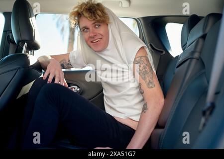 Ein lächelnder junger Mann mit Tätowierungen sitzt auf dem Rücksitz eines Autos, trägt einen weißen Hoodie und eine schwarze Hose. Stockfoto