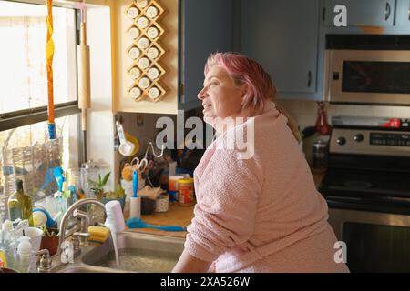 Eine lächelnde Frau in einem rosafarbenen Bademantel steht in einer sonnendurchfluteten Küche mit Hängeutensilien und einem Gewürzregal. Stockfoto