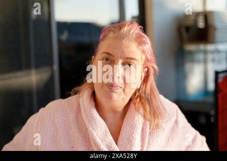 Frau mit rosafarbenen Haaren in einem gemütlichen rosa Bademantel, die drinnen sitzt und das Sonnenlicht ihr Gesicht beleuchtet Stockfoto