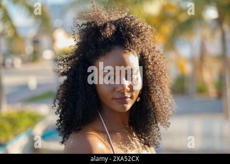 Nahaufnahme einer jungen Frau mit lockigen Haaren im Freien, in goldenem Sonnenlicht getaucht, die mit einem weichen Ausdruck direkt in die Kamera blickt. Stockfoto